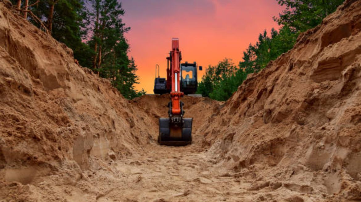 An excavator carves a trench in the midst of a forest, set against the breathtaking backdrop of a sunset. The backhoe is engaged in earthmoving operations, preparing the groundwork for the installation of pipelines for crude oil, natural gas, and water mains.